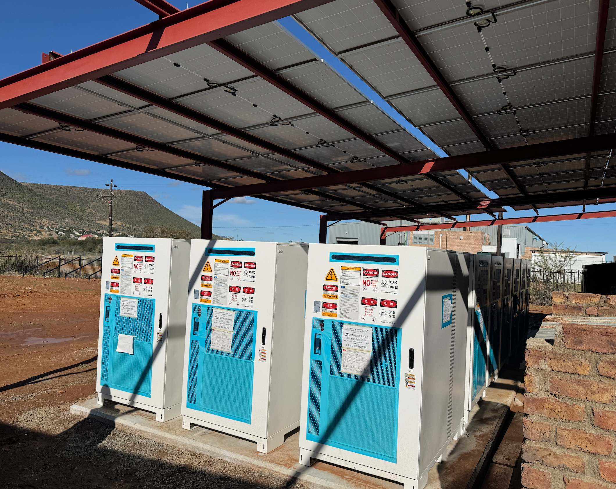 Phase 1 installation at Dr Beyers Naudé Municipality: battery energy storage cabinets mounted on concrete plinths beneath a solar PV carport canopy, with the Karoo hillside behind.